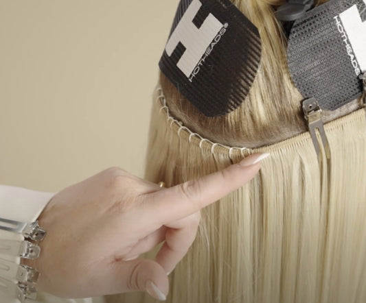 A close-up of hair extensions being applied, with a hand pointing at a row of sewn extensions. Hotheads-brand hair clips are used to section the hair.